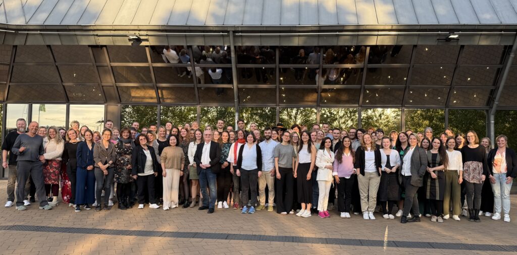 Gruppenfoto von über 100 Anwesenden (Mitarbeiter*innen, PiAs der Ambulanzen u. die 4 Leitungsgremiumsmitglieder) bei der Abschlussfeier-2025 auf der Terrasse der Fürther Stadthalle 